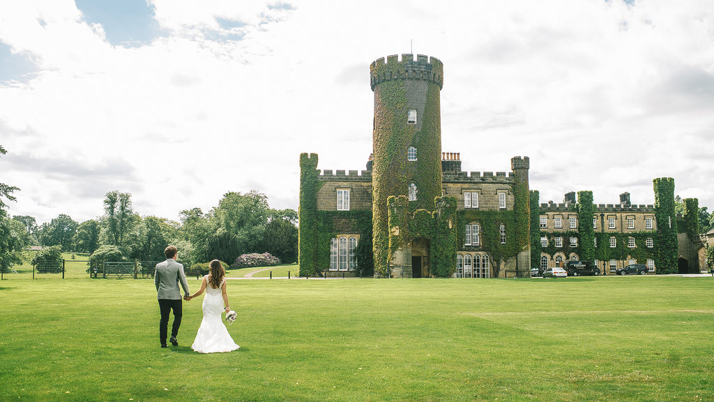 A bride and groom holding hands in front of the castle turret during a luxurious wedding at Swinton Park Hotel in North Yorkshire