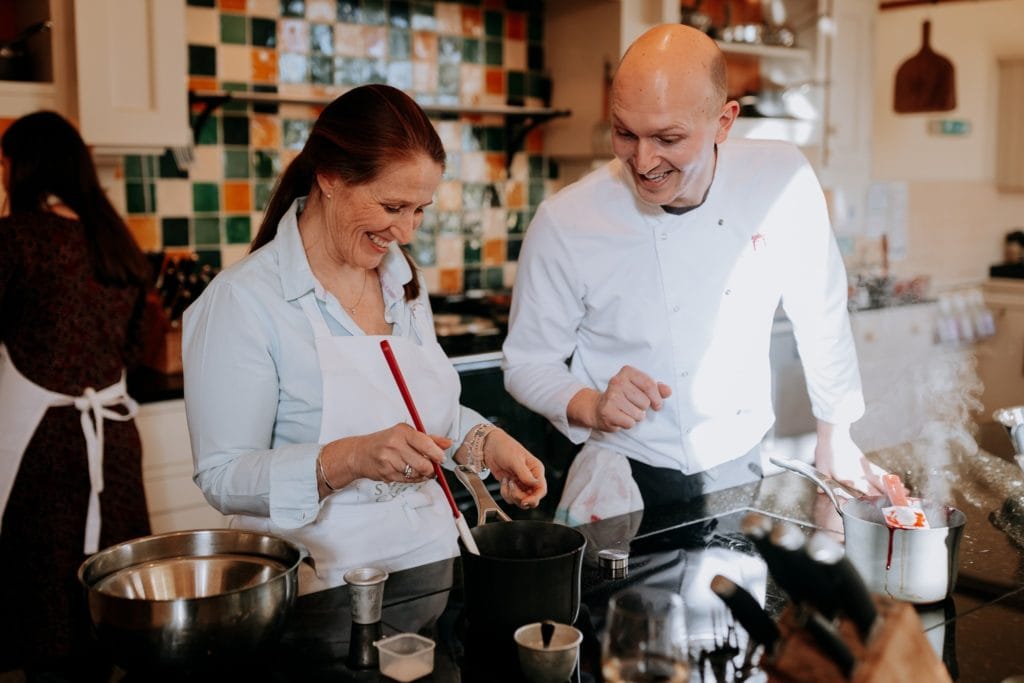 A smiling woman stirring a saucepan as a cookery course chef helps