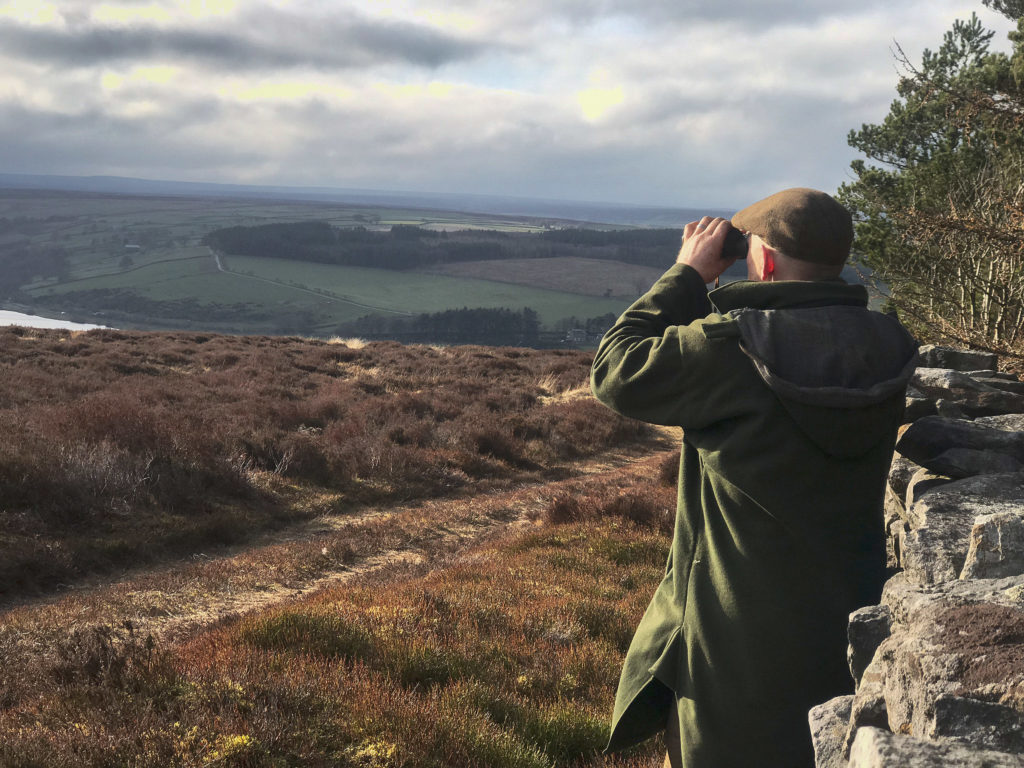 A man with binoculars birdwatching across the moors and Yorkshire countryside at Swinton Estate in North Yorkshire