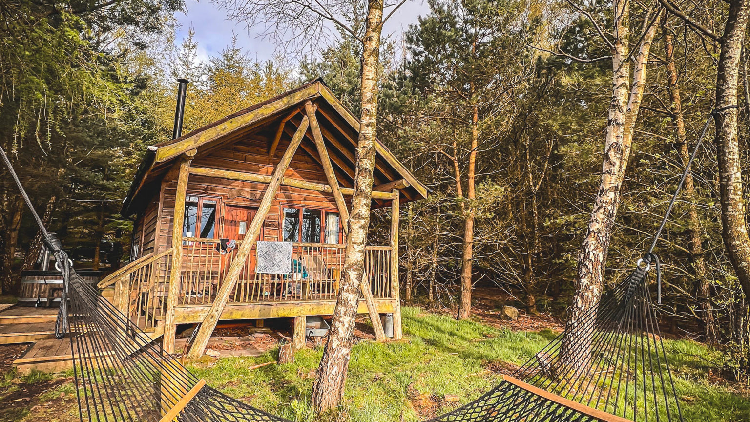 The exterior of a wooden Tree Lodge, with a hammock and woodfired hot tub, in the woodland at Swinton Bivouac in North Yorkshire