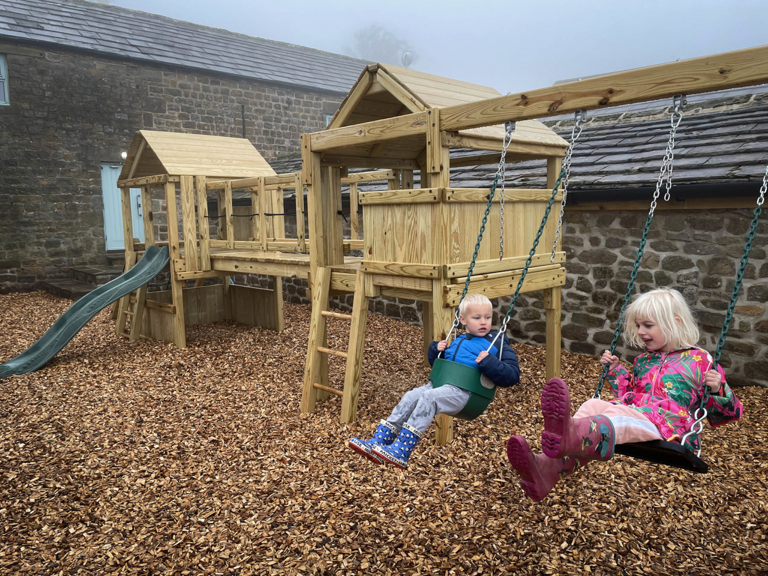 Two young children wearing coats playing on swings at the Swinton Bivouac play area
