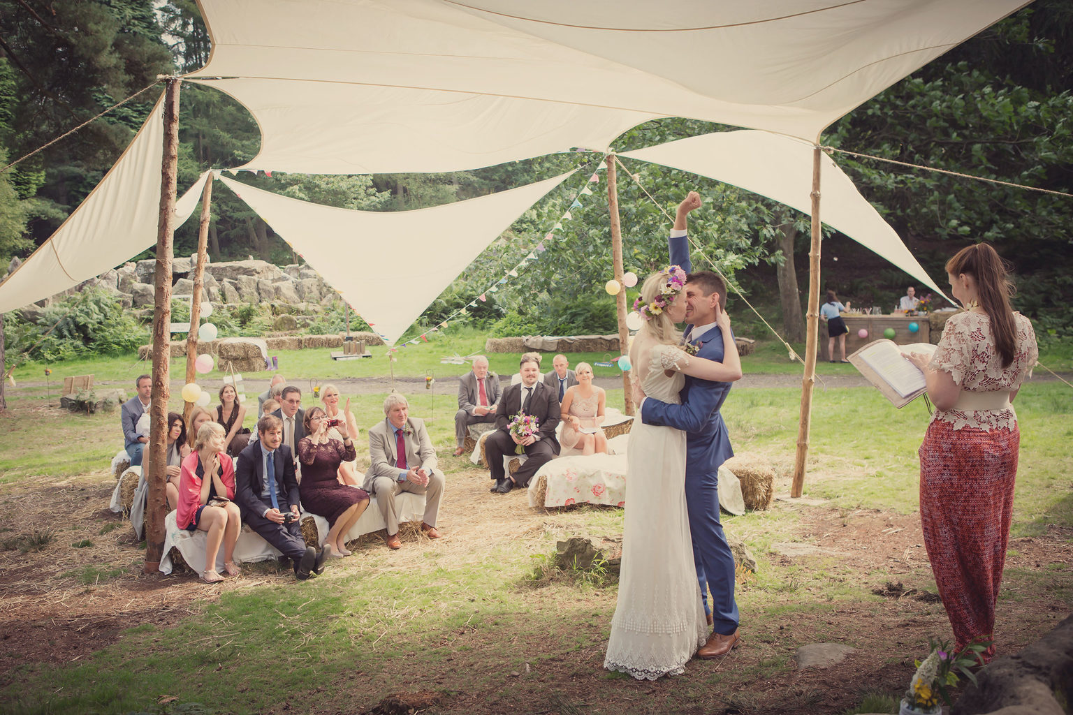 A bride and groom kissing during an outdoor boho wedding ceremony in the woodland at Swinton Bivouac in North Yorkshire. Photo by Lissa Alexander.